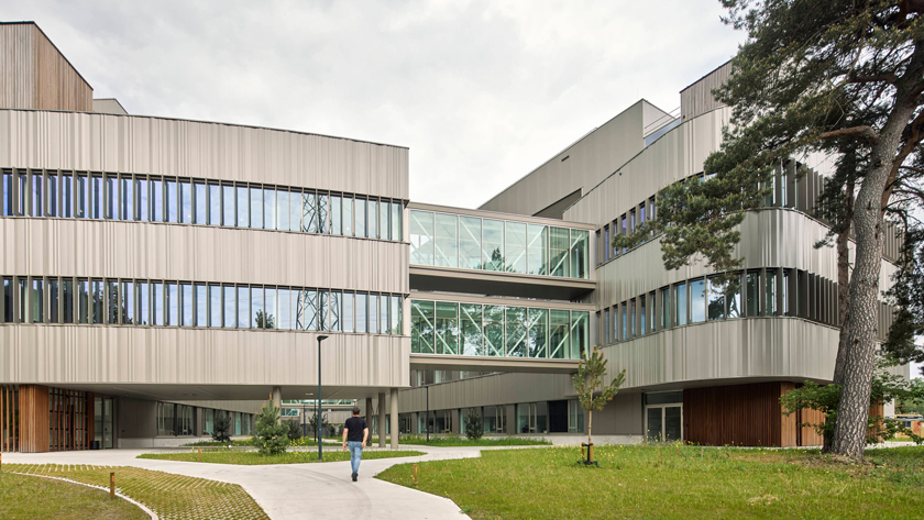Modern multi-story campus building with glass skywalk and a person walking along a curved path on a grassy courtyard.