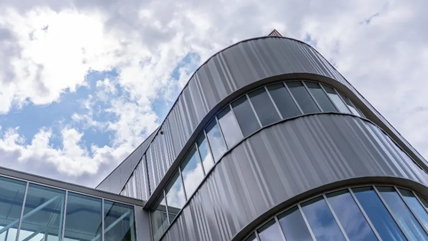 Curved metal-clad building with glass windows reaching toward a blue sky with clouds.