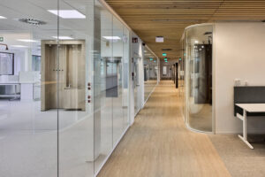 Long glass-walled office hallway with curved glass doorway on the right and wooden ceiling slats.