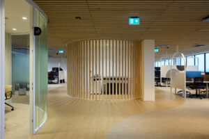 Open office area with a circular wooden slat partition, light wood flooring, and workstations beyond, creating a modern, airy workspace