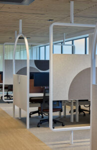 Open-plan office with fabric-panel partitions on white curved frames, desks, and rolling chairs near large windows at the back.
