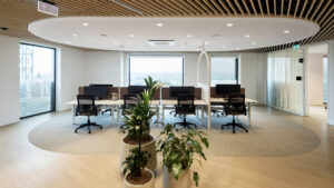 Open-plan office with rows of desks, computers, black chairs, and a circular white ceiling feature.