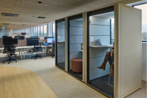 Open-office layout with rows of desks and glass-walled private pods along a window wall; a person sits inside one pod wearing brown boots.