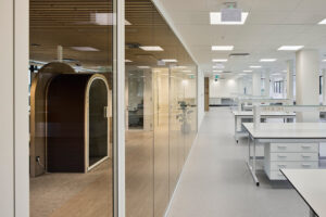 Open-plan office with glass-walled rooms, a wooden pod in the foreground, and bright white desks behind it.