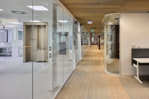 Glass-walled office corridor with a wooden slat ceiling and a curved glass meeting room on the right.