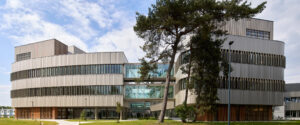 Modern curved beige building with glass walls and vertical slats, set in a park-like foreground with trees and a blue sky.