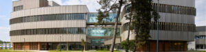 Modern curved office/campus building with beige panels, large glass windows, and trees out front on a sunny day.