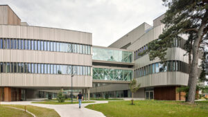 Modern multi-story campus building with glass skywalk and a person walking along a curved path on a grassy courtyard.