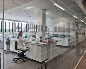 Scientist in a white lab coat and gloves sits at a benchtop in a modern laboratory, performing an experiment with pipettes and equipment behind a glass wall with vertical stripes.