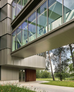 Contemporary building with a glass-enclosed elevated walkway extending from the façade, metal panels, and surrounding greenery.