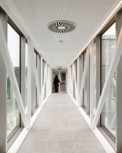 Long indoor walkway with white diagonal supports, large glass windows, and a person walking away at the end of the corridor