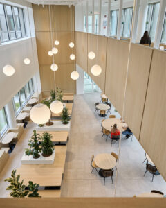 Bright, open stairwell-like atrium with large globe pendant lights, light wood panels, and seating areas with plants and round tables nearby