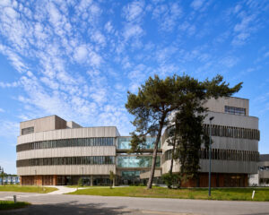 Modern curved office building with white metal cladding, long horizontal windows, and a tall pine tree in front. The blue sky is clear above.