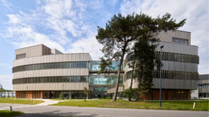 Modern multi-story office building with curved light-gray panels and glass atrium, surrounded by trees. Refrains from describing people or weather beyond setting.