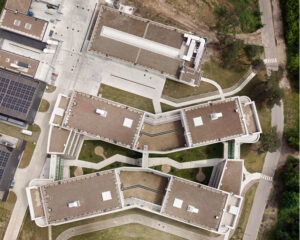 Aerial view of a campus-style building complex with interconnected flat-roofed structures, walkways and green lawns, viewed from above.