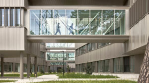 Modern office courtyard with a glass pedestrian bridge connecting two buildings; silhouettes walk inside the bridge above a landscaped area.