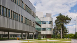 Modern gray metal building with a curved glass facade and a person walking beneath it on a sunny day.