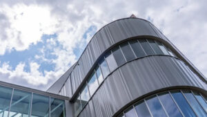 Curved metal-clad building with glass windows reaching toward a blue sky with clouds.